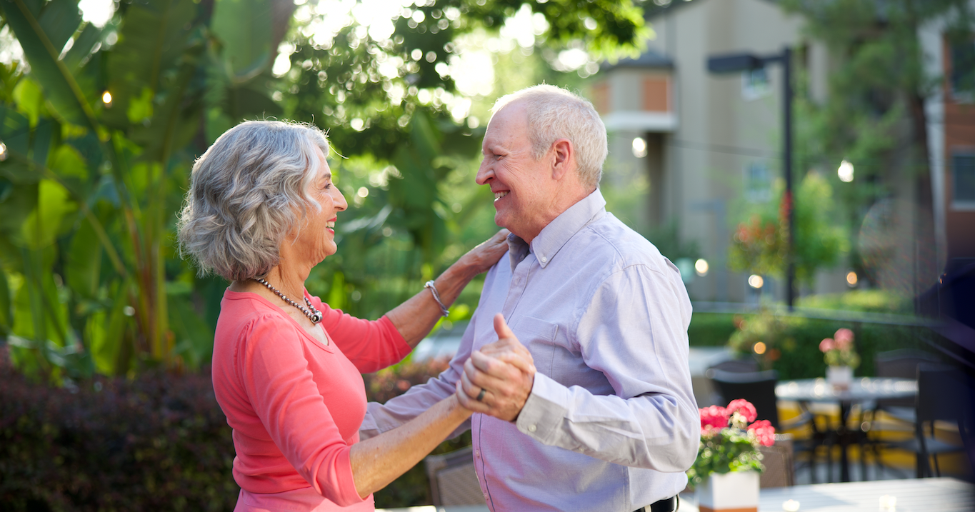 An older couple smiles joyfully while dancing together outdoors in a sunlit garden setting, surrounded by greenery and flowers.